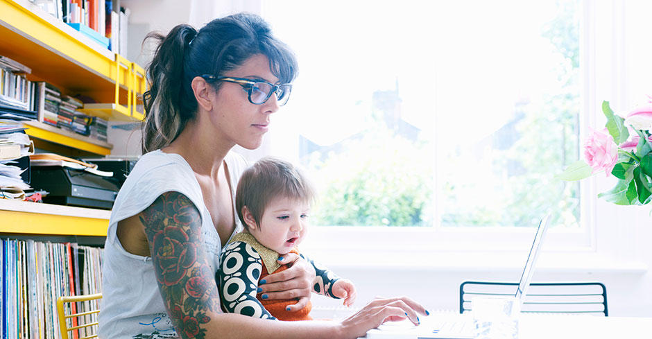 Tatooed woman holds her infant while doing laptop work in an office